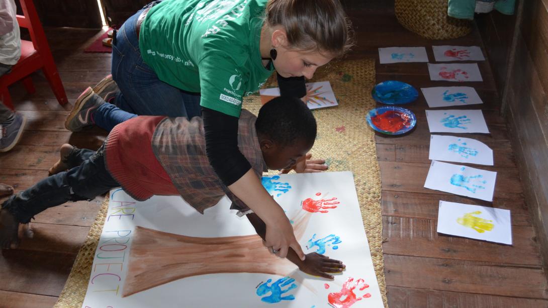 A volunteer on a mission trip to Madagascar, Africa helps a child with painting a tree. On a mission trip to Africa for high school students, a volunteer helps a child on an art project.